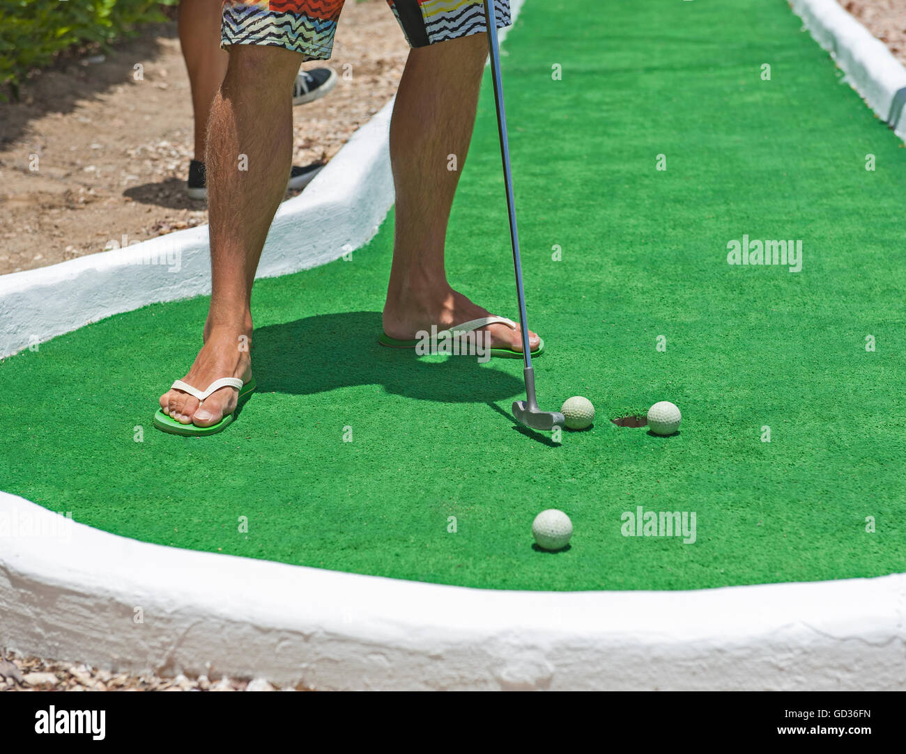 Man in flip flop sandals on summer vacation playing mini golf putting ball at hole Stock Photo