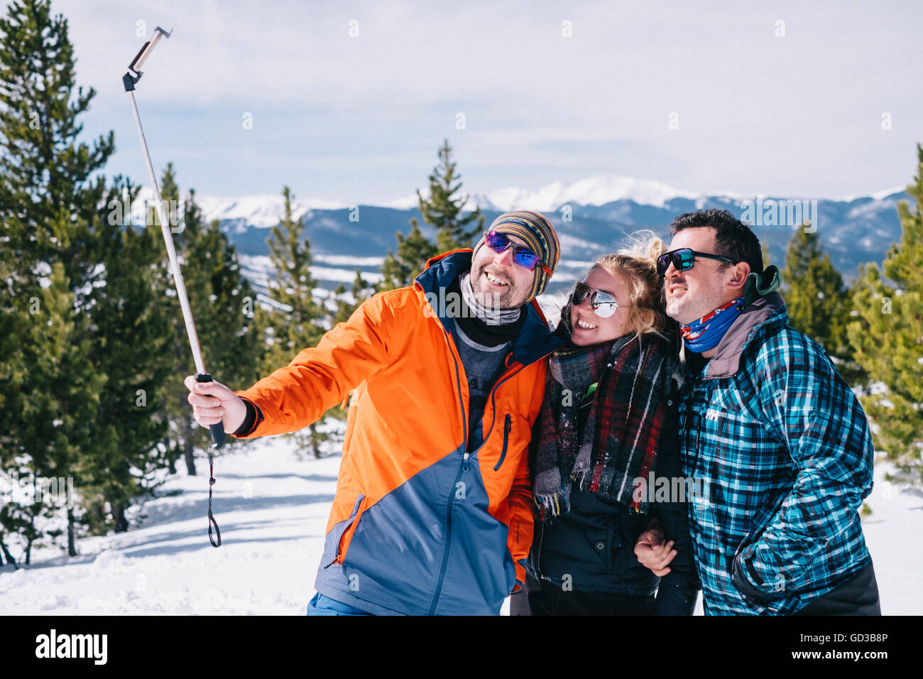 Three people, two men and a young woman in skiing gear posing for a selfie, one holding a selfie stick. Stock Photo