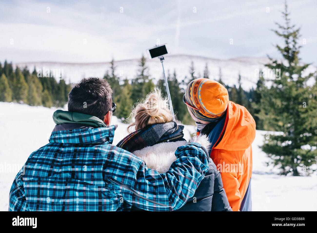 Three people, two men and a young woman in skiing gear posing for a selfie, one holding a selfie stick. Stock Photo