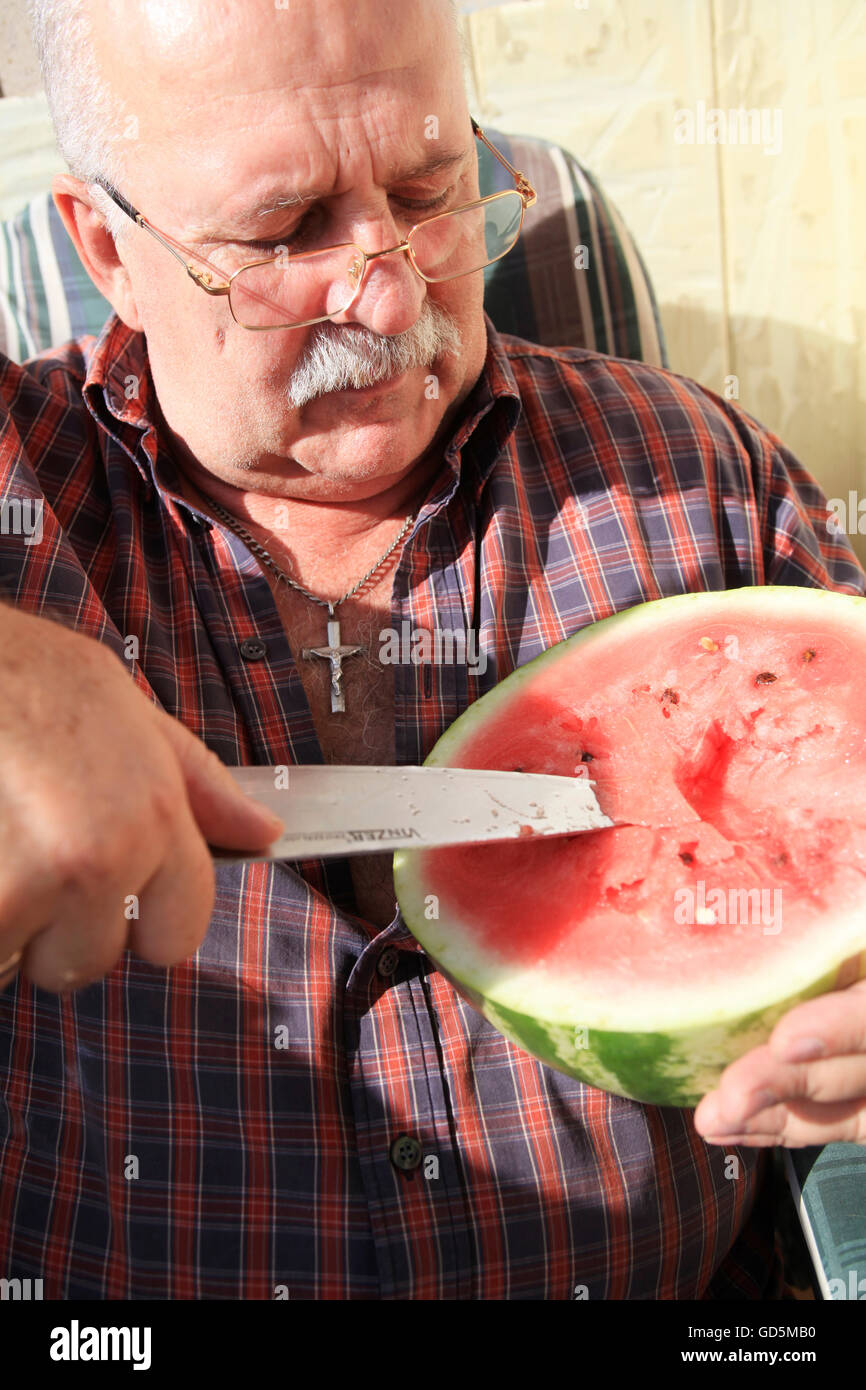 Ukrainian man of some 55-60 is cutting a water melon with a big knife, enjoying rich Ukrainian fruits and vegetables harvest Stock Photo