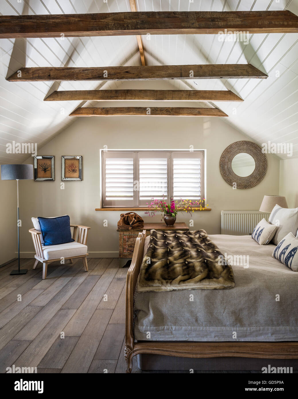 Plantation shutters and white wooden ceiling cladding in attic bedroom with vintage french cane bed. the chair is by Pomax Stock Photo