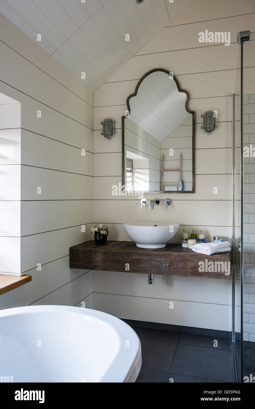 Basin resting on slab of timber in bathroom with walls lined white painted boards and a vintage mirror Stock Photo