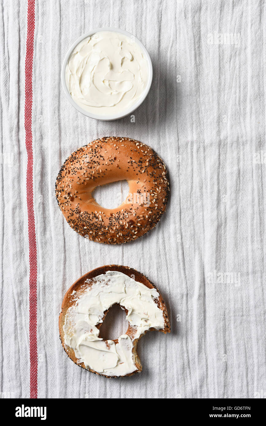 Overhead view of a sliced bagel, one half spread with cream cheese. A crock full of spread on a kitchen towel. Stock Photo