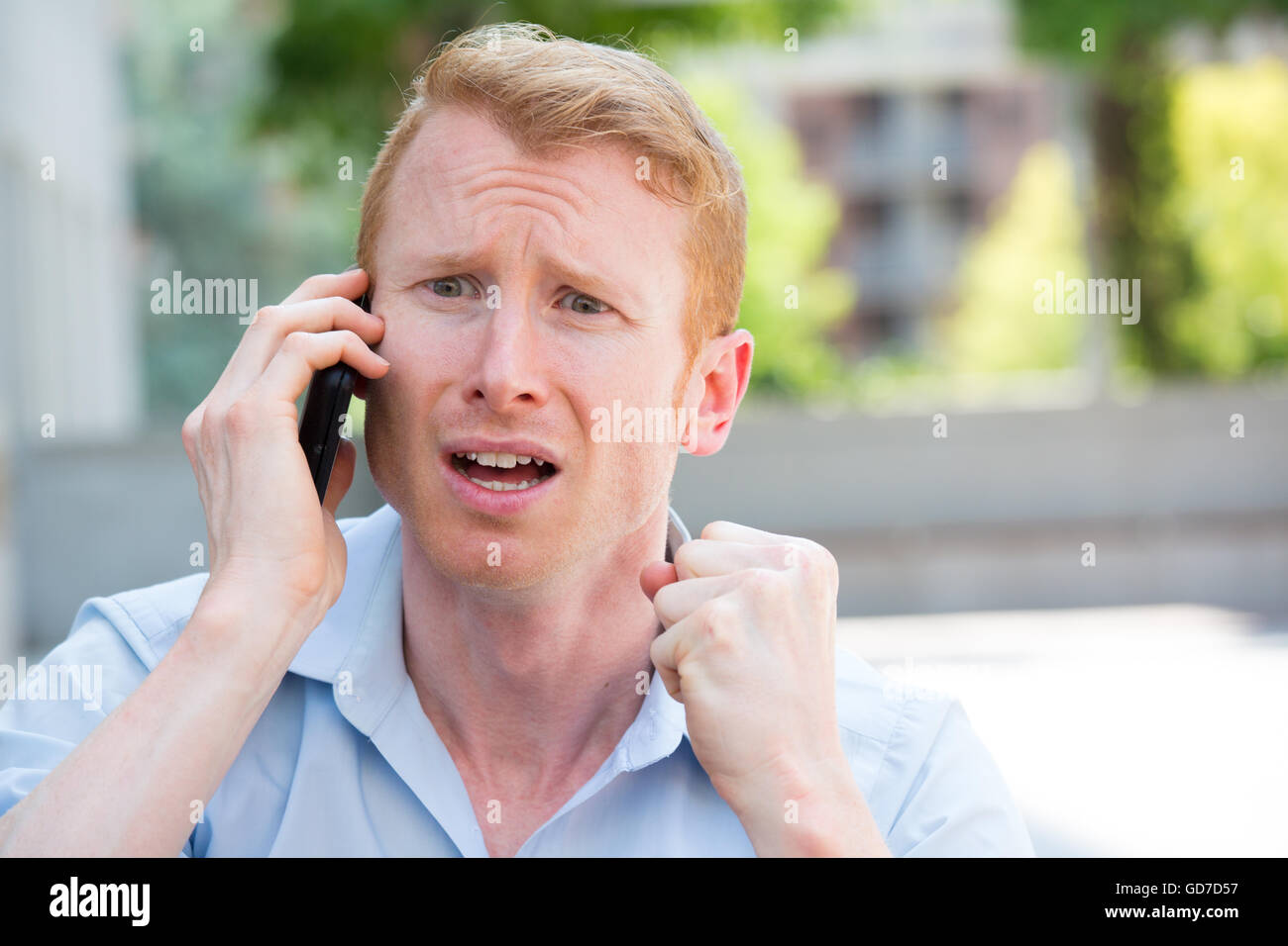 Closeup portrait, worried young man in blue shirt talking on phone to someone, looking gloomy, isolated outdoors outside backgro Stock Photo