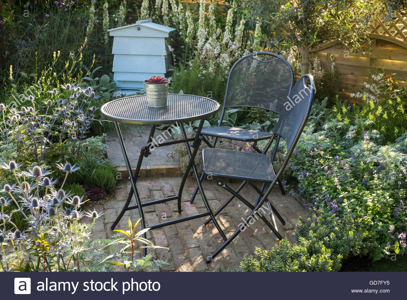 Small Summer Cottage Style Garden With Brick Patio, Metal Table And Small summer cottage style garden with brick patio, metal table and chairs, and silver white and blue planting. - Small Summer Cottage Style Garden With Brick Patio, Metal Table And