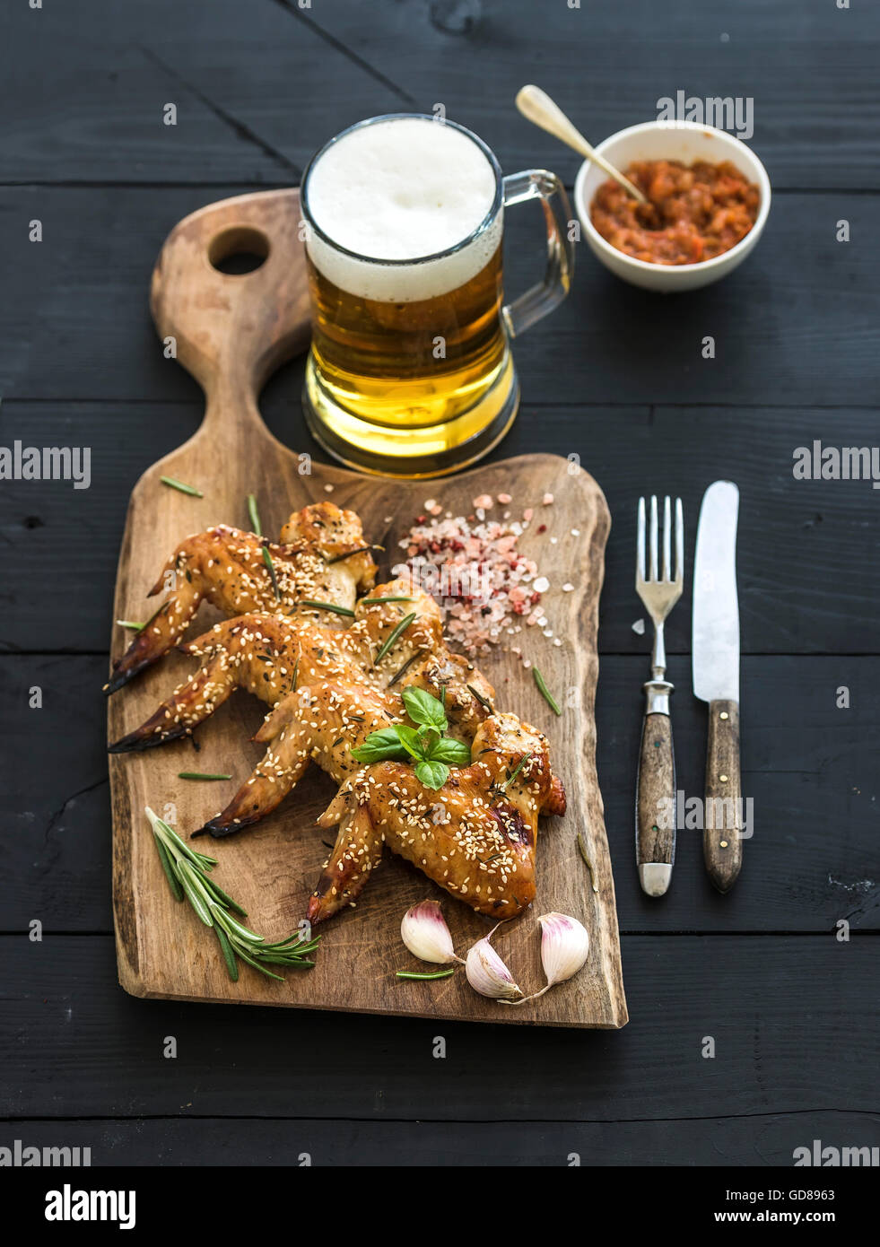 Grilled chicken wings with seasam, spices and herbs, glass of beer and tomato sauce on rustic wooden board over dark wooden back Stock Photo