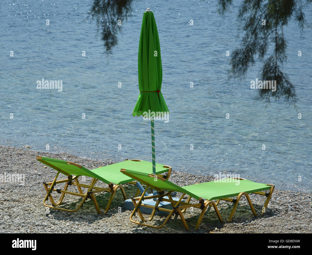 Two empty lime green sun loungers with a closed parasol sun shade on a shingle beach by the water's edge Stock Photo