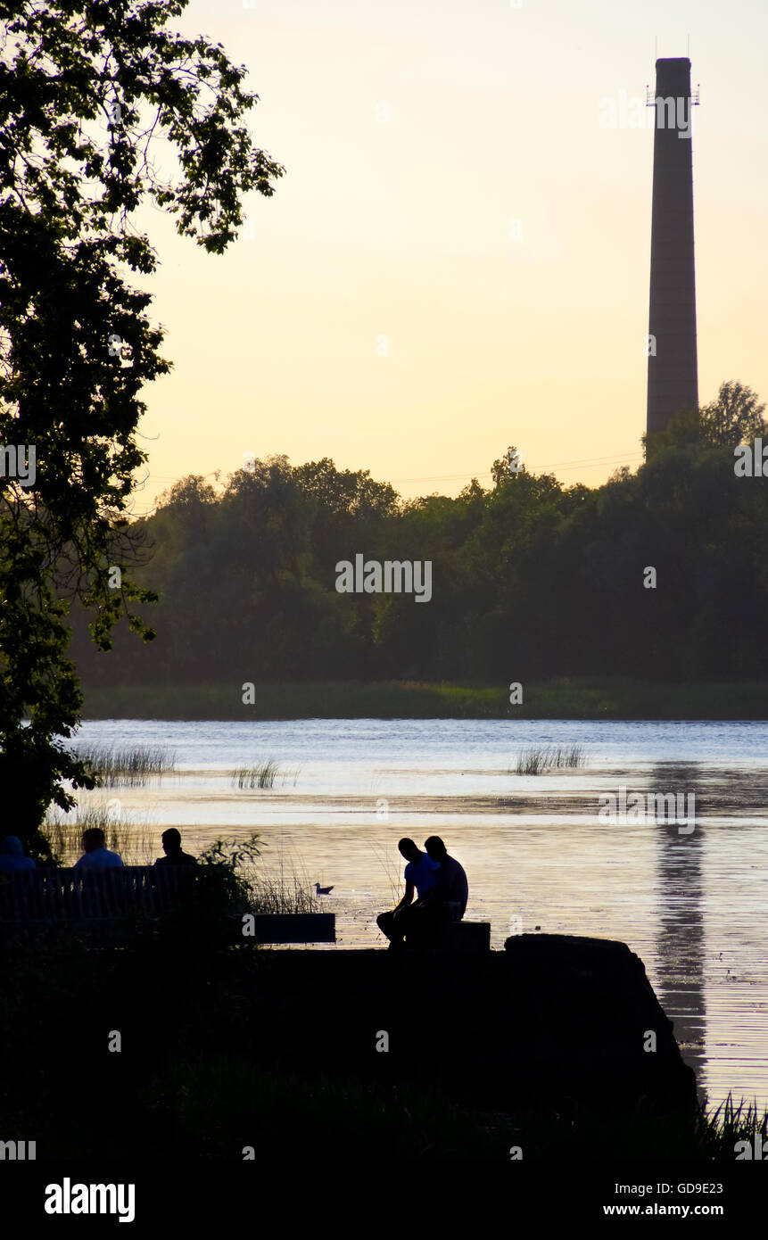 People sitting by the Pärnu River on a tender summer evening with the distant factory chimney reflecting in the water Stock Photo