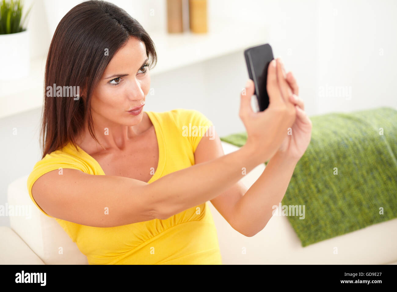 Attractive young woman in yellow shirt taking a selfie while sitting indoors Stock Photo