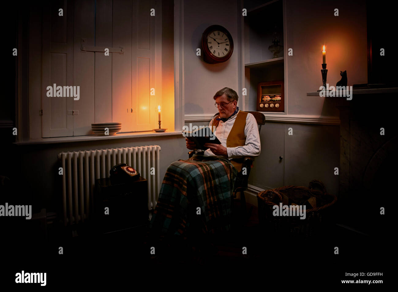 Older man sitting reading a book by candlelight with valve radio on and rug on knees Stock Photo