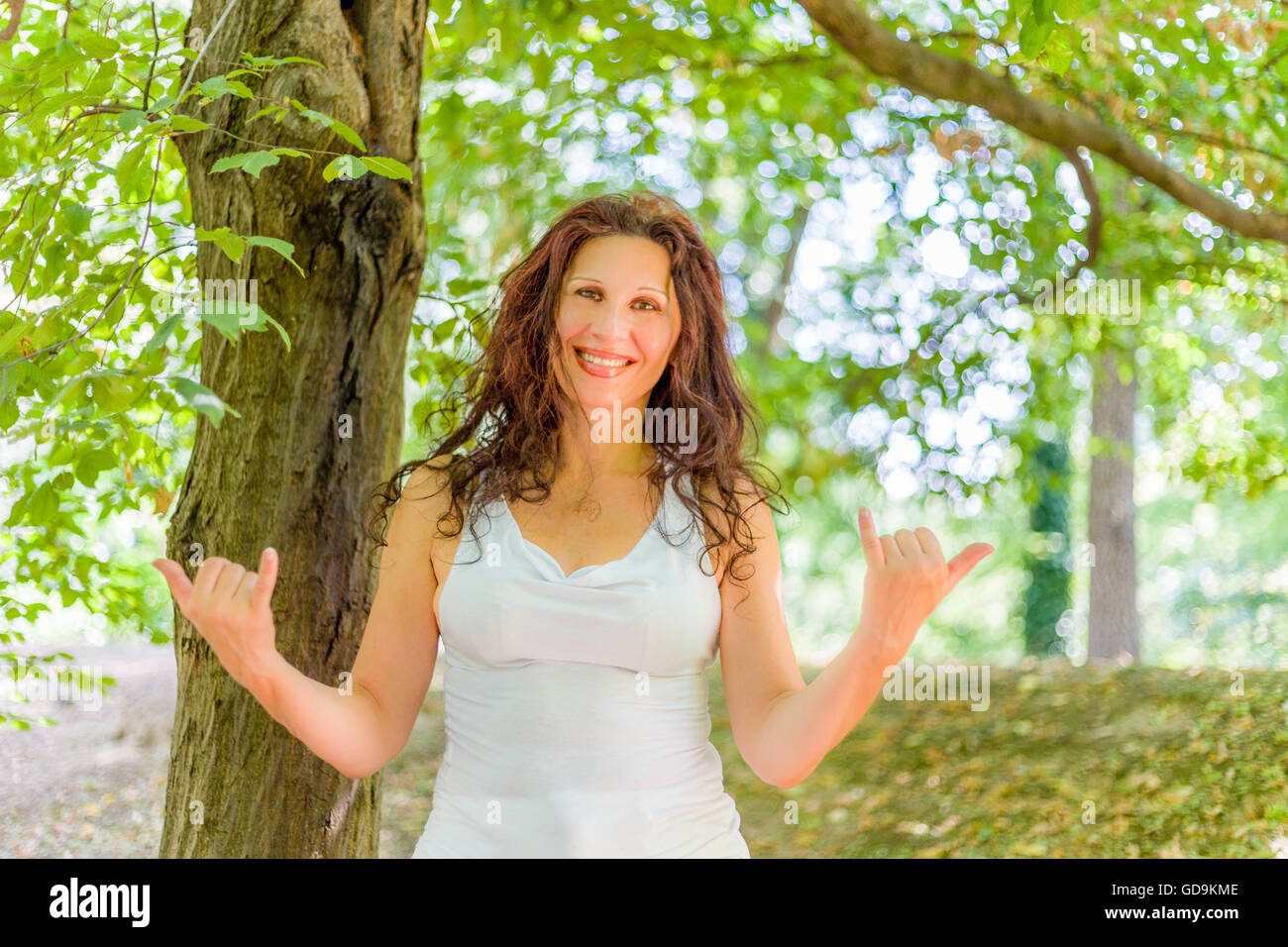 Close up of busty classy mature woman smiling at the camera while doing welcome shaka sign against green garden background with copy space Stock Photo