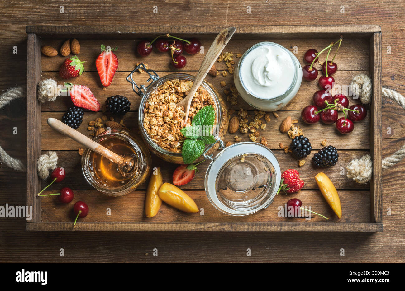 Healthy breakfast ingredients. Oat granola in open glass jar, fruit, yogurt, honey, berries and mint in wooden tray over wooden Stock Photo