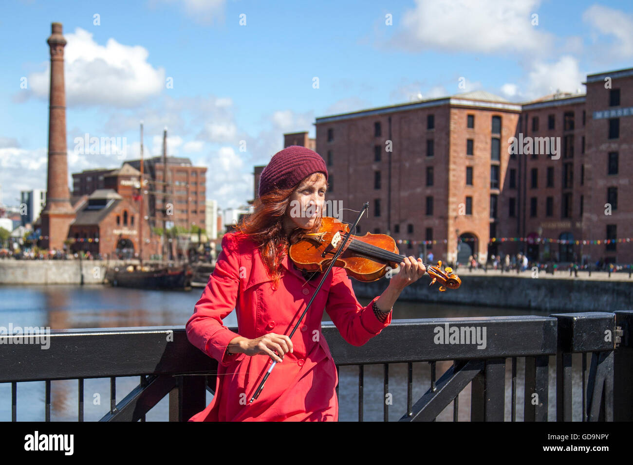 Female street busker playing the violin or fiddle with Albert dock in the background, in dockland Liverpool. Buskers on the coastal riverside promenade on Liverpool’s Waterfront, Merseyside, UK.  The Pier Head is a central major tourist attraction with several notable landmarks and modern developments which focus attention on the cities long history and heritage as a major port on the Mersey. Stock Photo