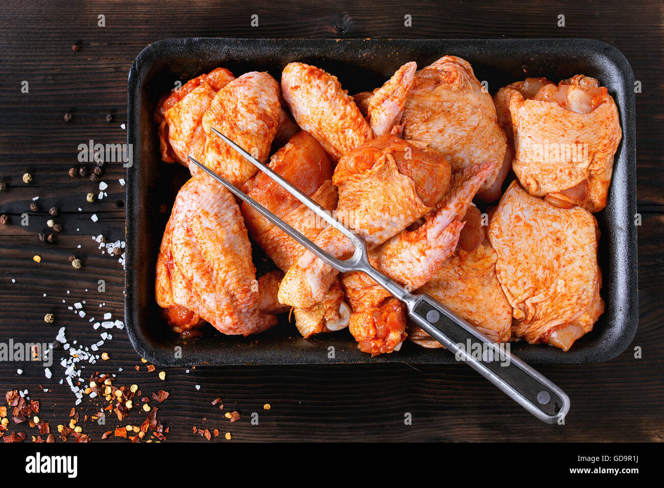 Raw Marinated chicken meat wings and legs for BBQ, served in plastic box with seasoning and vintage meat fork over dark wooden b Stock Photo