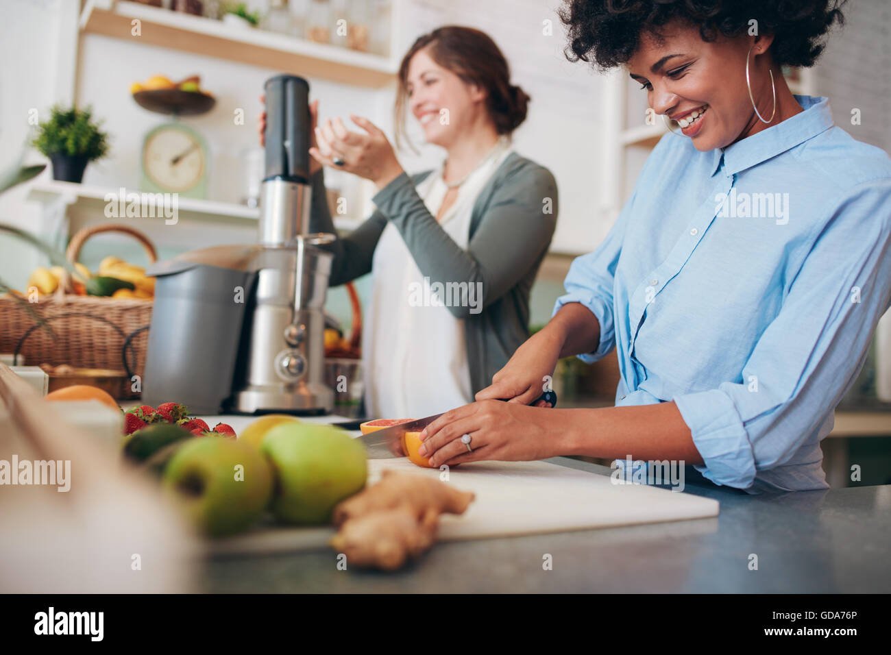 Young african woman cutting fruits at bar counter, with coworker using juicer in background. Two female working at juice bar. Stock Photo