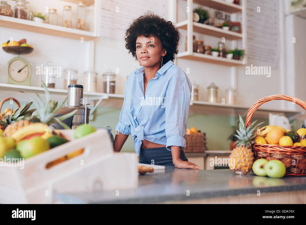 Portrait of attractive young woman standing at juice bar counter. African female employee at juice bar looking away. Stock Photo