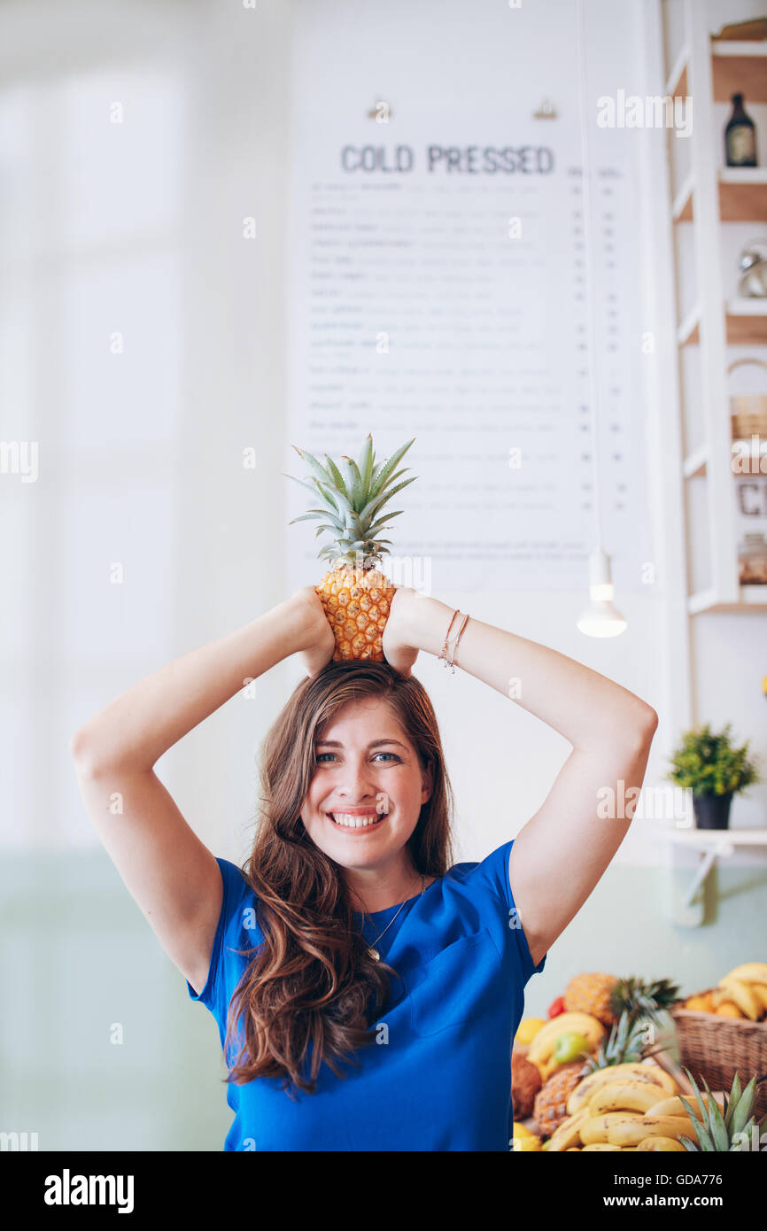 Portrait of happy young woman holding a pineapple on her head. Beautiful caucasian female at juice bar. Stock Photo
