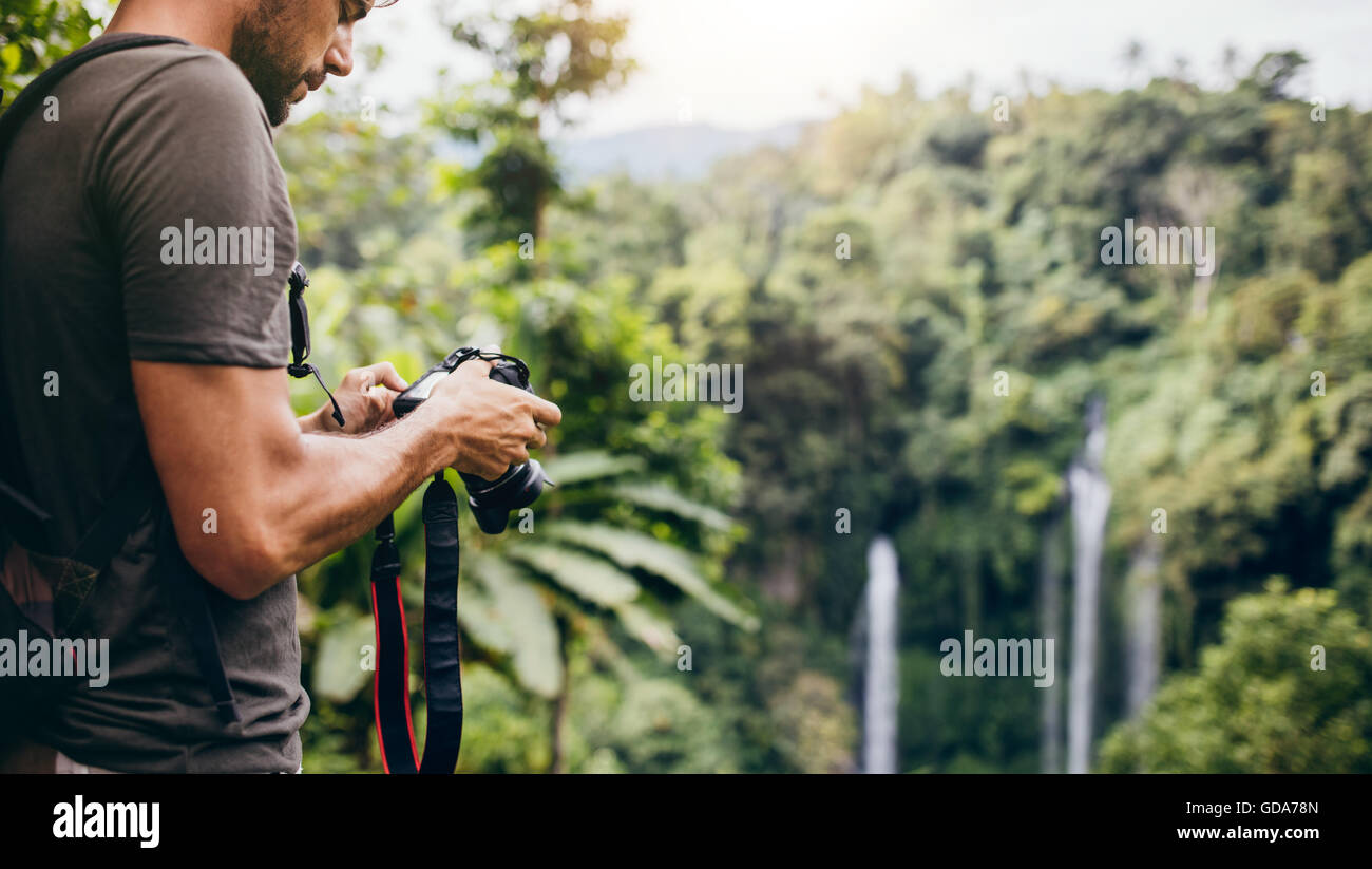 Young man looking at his camera while standing by a waterfall in forest. Male hiker standing on a cliff and looking the waterfal Stock Photo