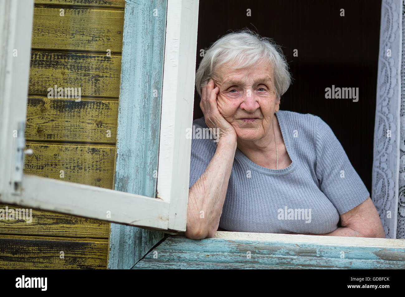Elderly woman pensioner looks out the window of a village house. Stock Photo