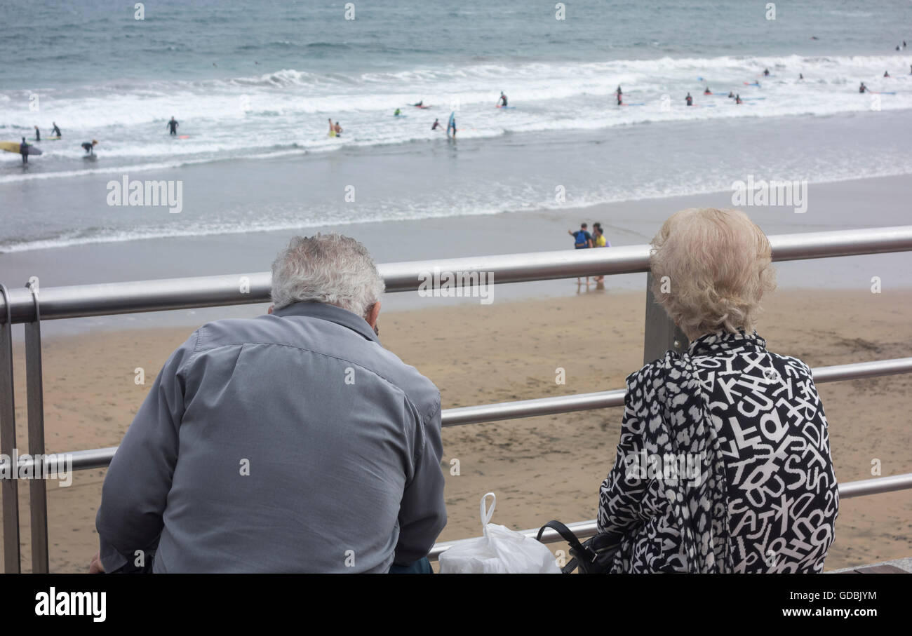 Elderly couple watching surfers on beach. Stock Photo