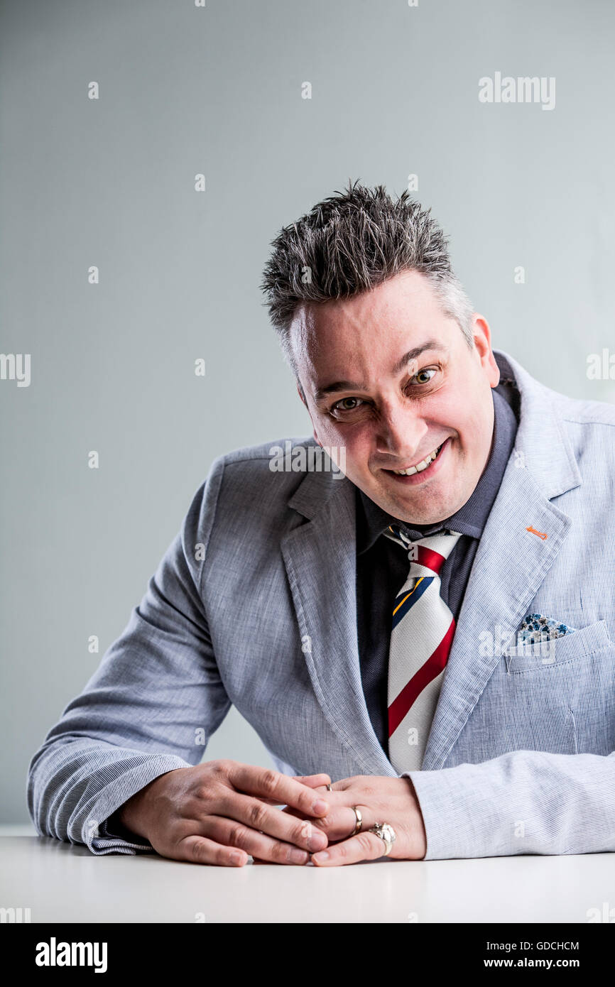 gray hair man in suit jacket look at the camera with a dodgy unreliable face and hands on the desk. Stock Photo