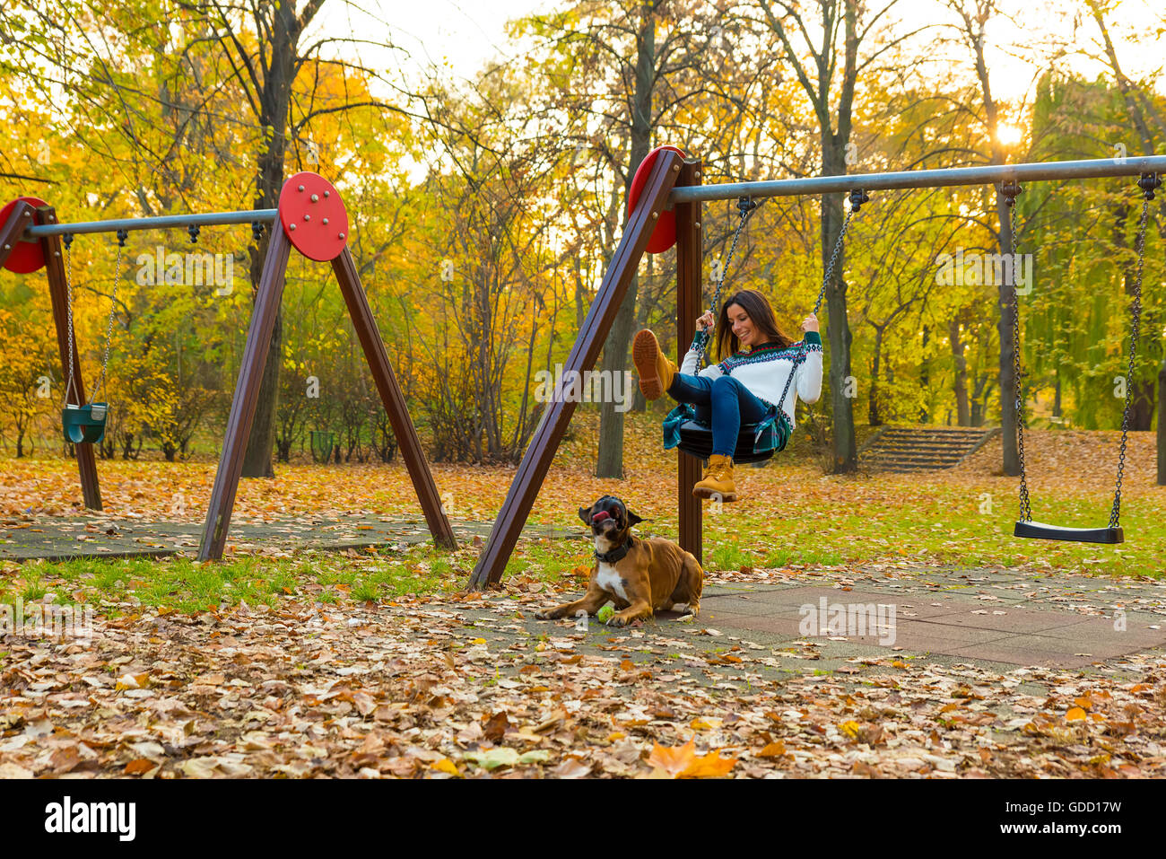 Beautiful young woman enjoying the autumn in the forest on a swing with her dog around. Stock Photo
