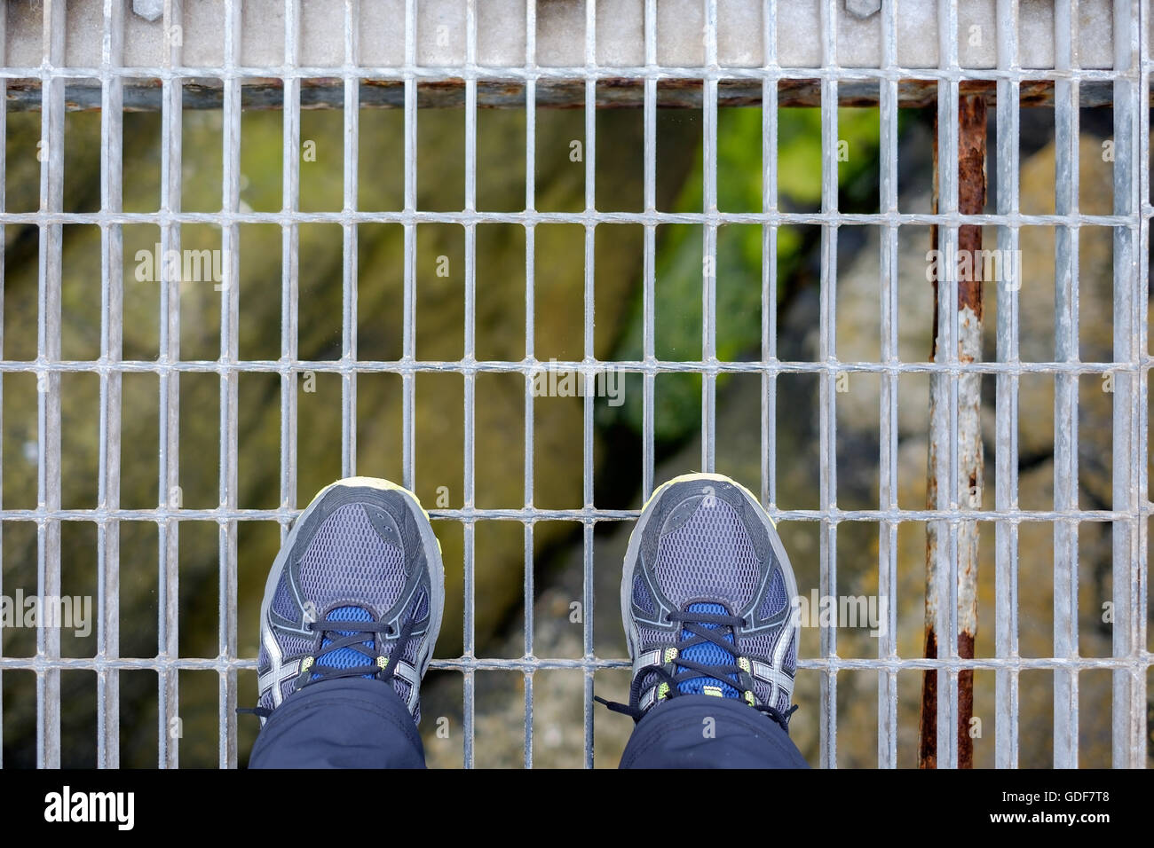 Two feet standing on a metal grate looking down to rocks below Stock