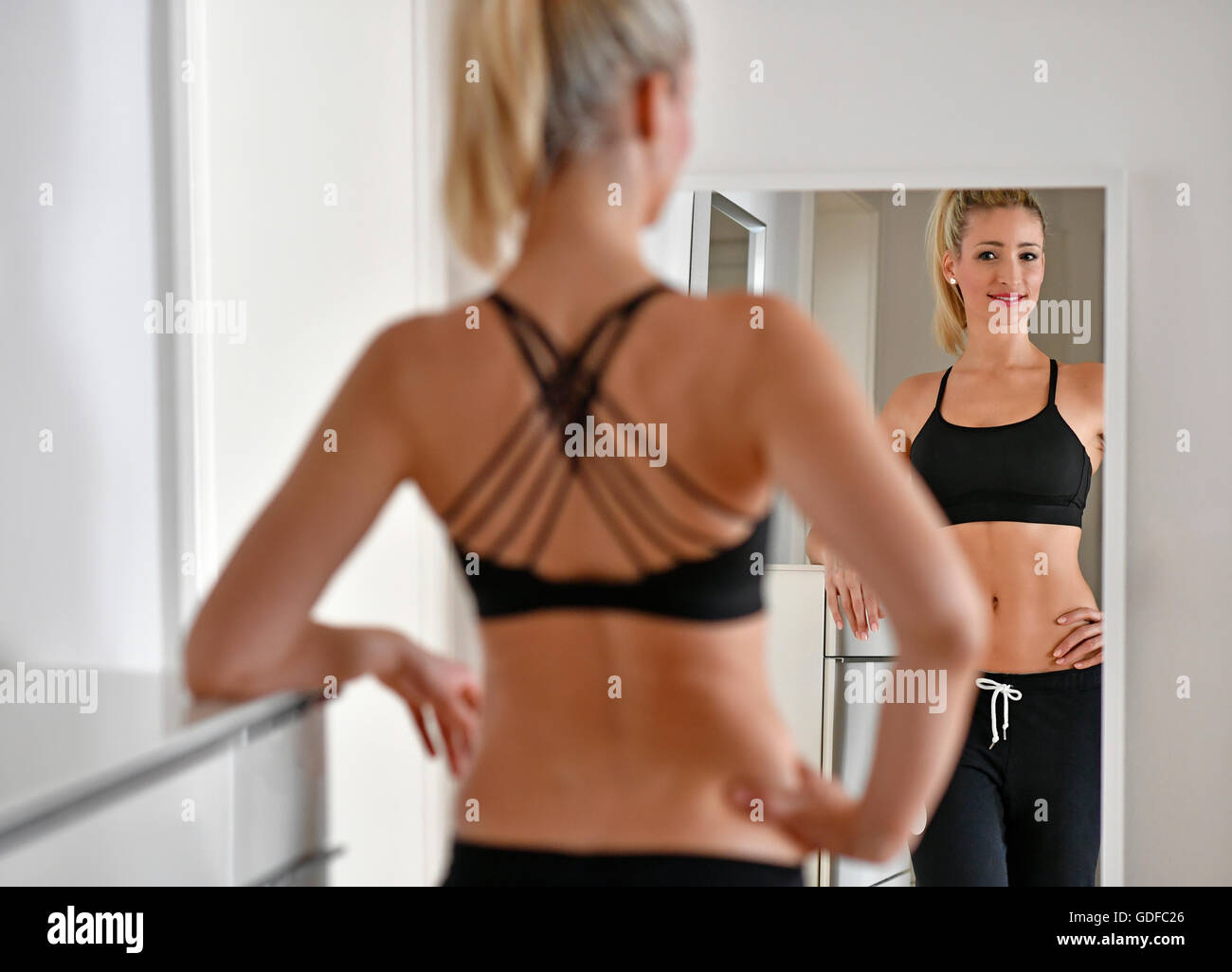 Young woman with sports bra and sweatpants looks at herself in the mirror Stock Photo