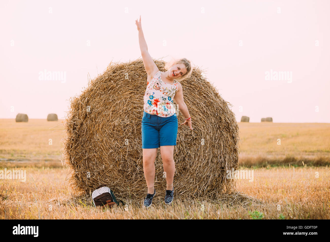 Beautiful Plus Size Young Woman Girl Jumping Near Haystack With His Hand Up And Smiling. Sunset Of The Day In Summer Field Meado Stock Photo