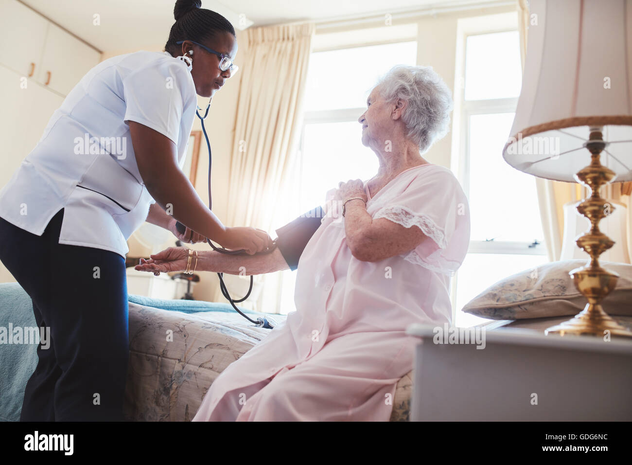 Female nurse checking blood pressure of a senior woman sitting on bed at home. Home carer checking patients blood pressure at ho Stock Photo