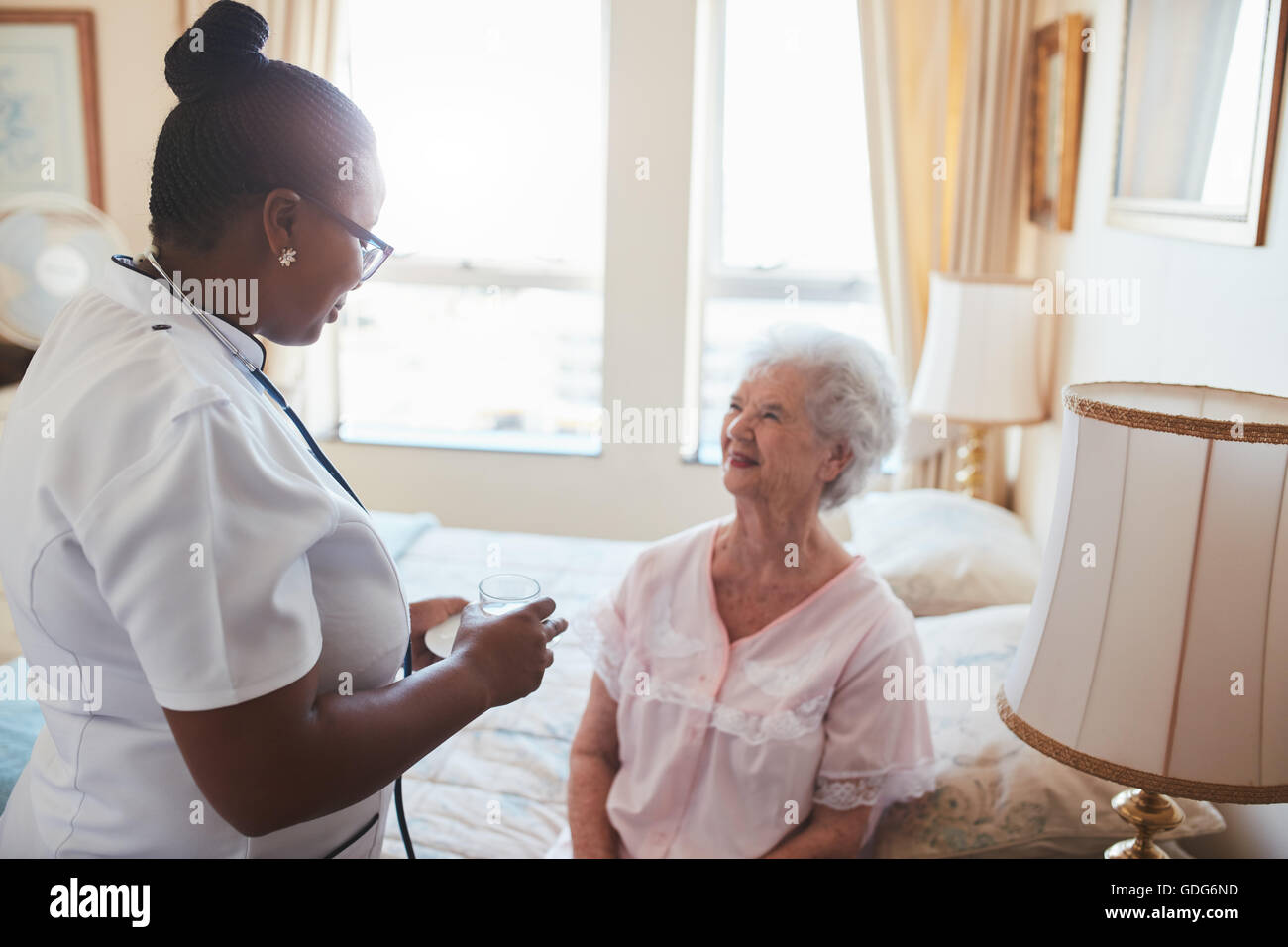 Female nurse giving medicine to senior female patient at home. Elderly woman sitting on bed with home caregiver standing by with Stock Photo