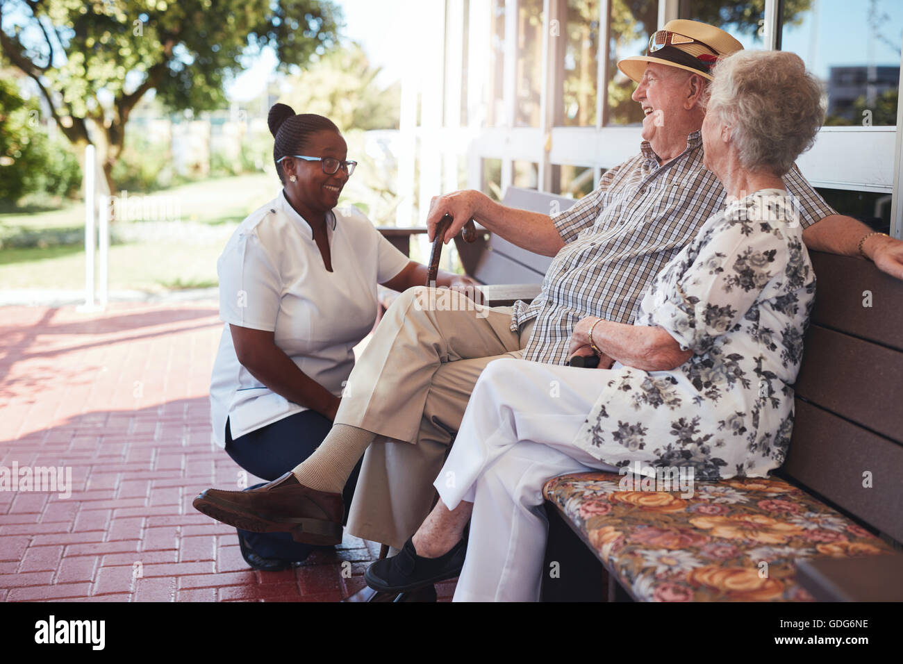 Portrait of loving retired couple relaxing on a bench outside their house with a female nurse sitting by. Caucasian elderly man Stock Photo
