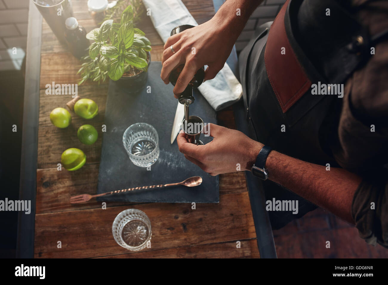 Close up of barman hands pouring alcoholic drink in to a jigger to prepare a cocktail, with basil leaves and lemons on the bar c Stock Photo