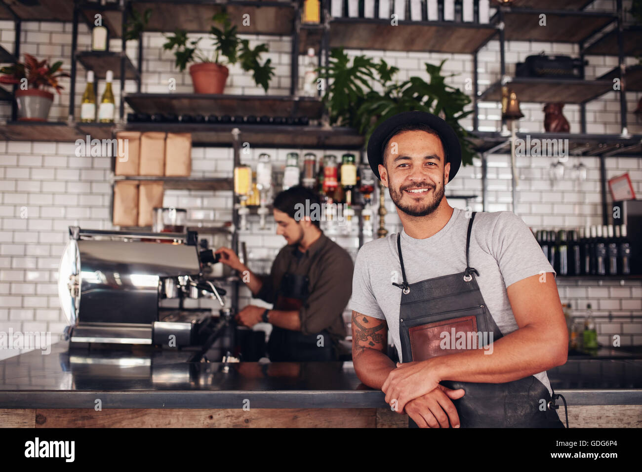 Portrait of male coffee shop owner standing at the counter with barista working in background making drinks. Stock Photo