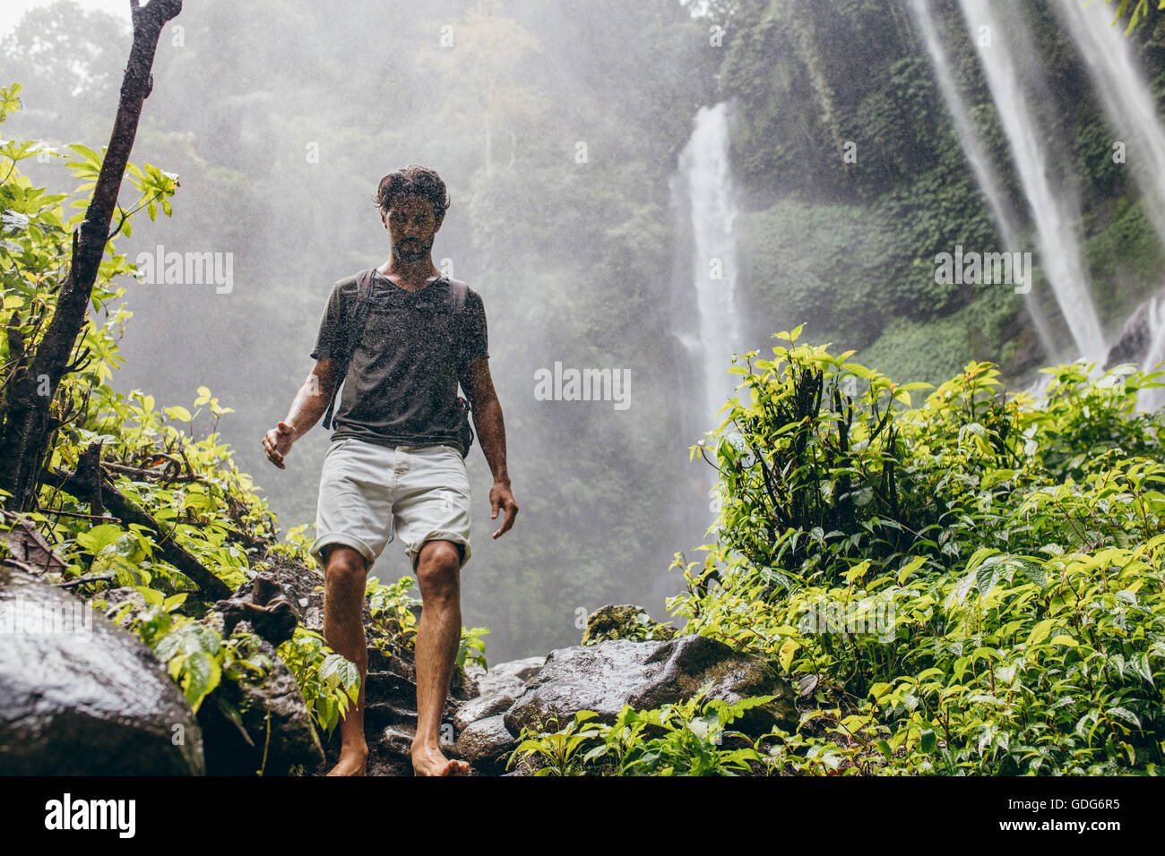 Outdoors shot of caucasian male hiker walking down the mountain trail. Young man hiking in forest with waterfall in background. Stock Photo