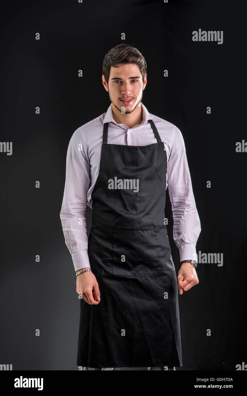 Young chef or waiter posing, wearing black apron and shirt, on dark background Stock Photo