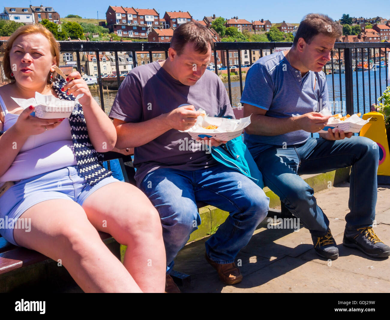 Two men and a woman sitting by Whitby Harbour enjoying a meal of takeaway Fish and Chips Stock Photo