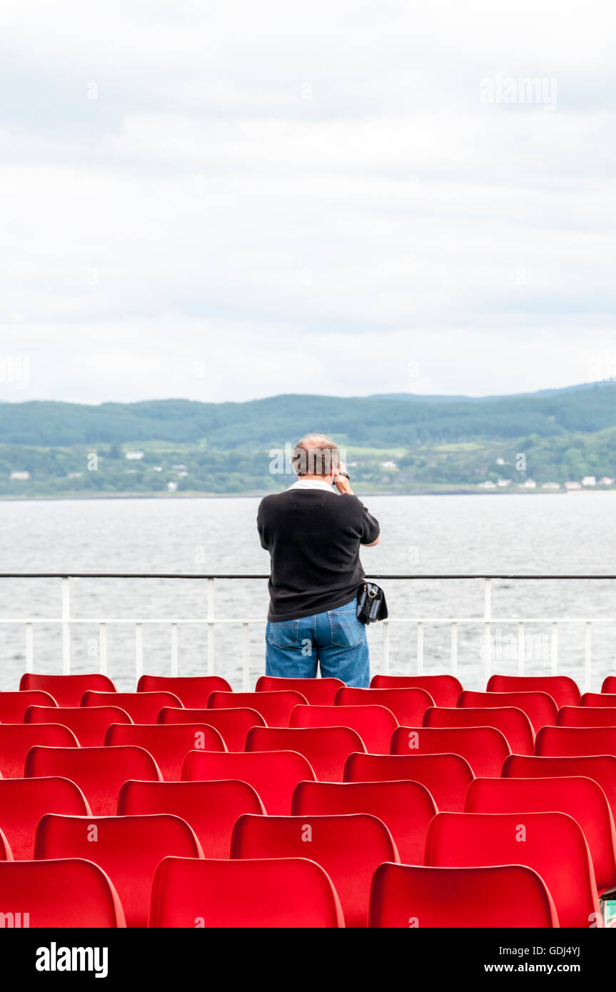 Man taking photographs or video of the shore of mainland Scotland at Oban from stern of ferry to Isle of Barra. Stock Photo