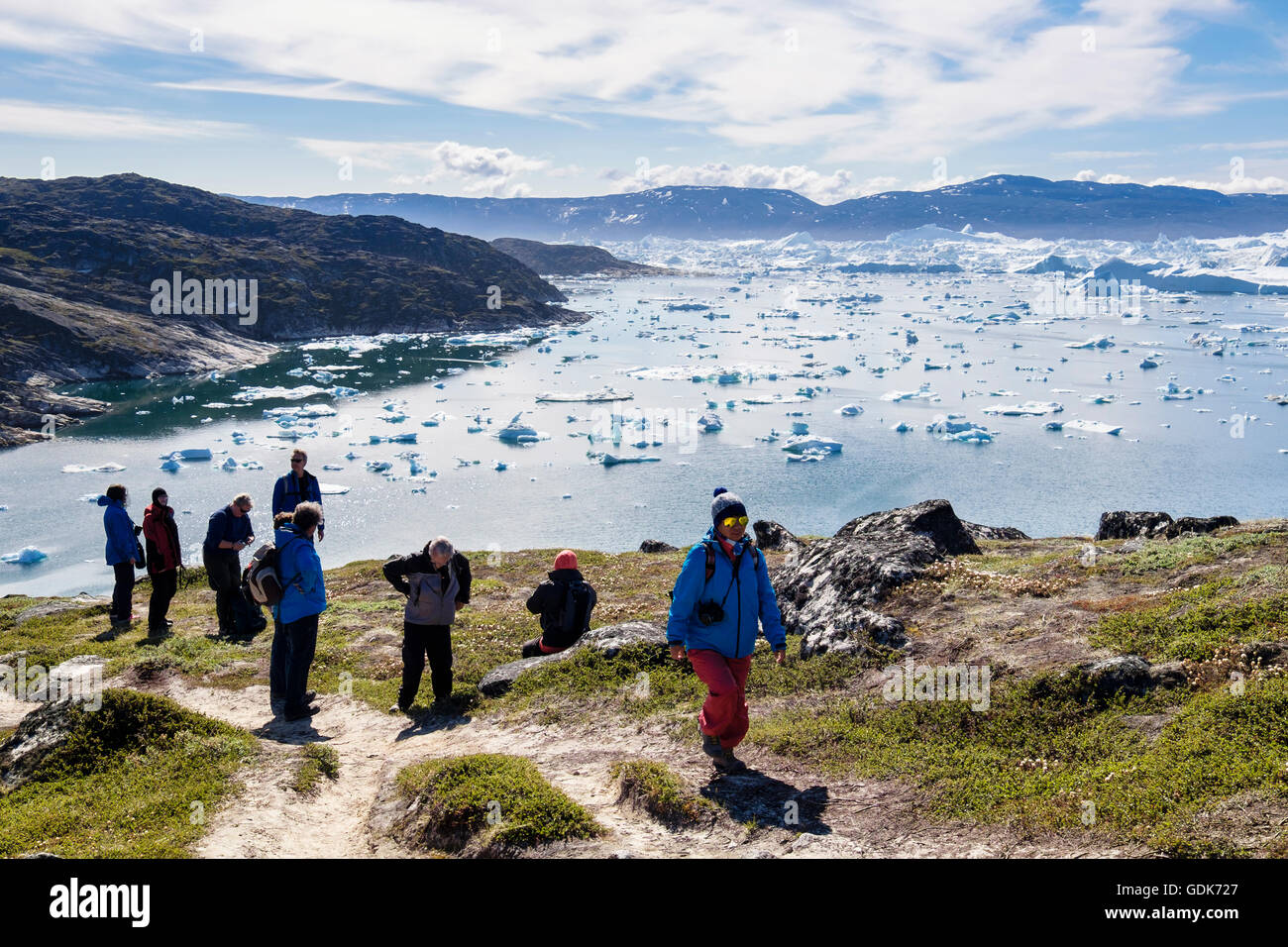 Hikers hiking on blue hike trail path by Jakobshavn or Ilulissat Icefjord with large icebergs in fjord in arctic summer. Ilulissat Western Greenland Stock Photo