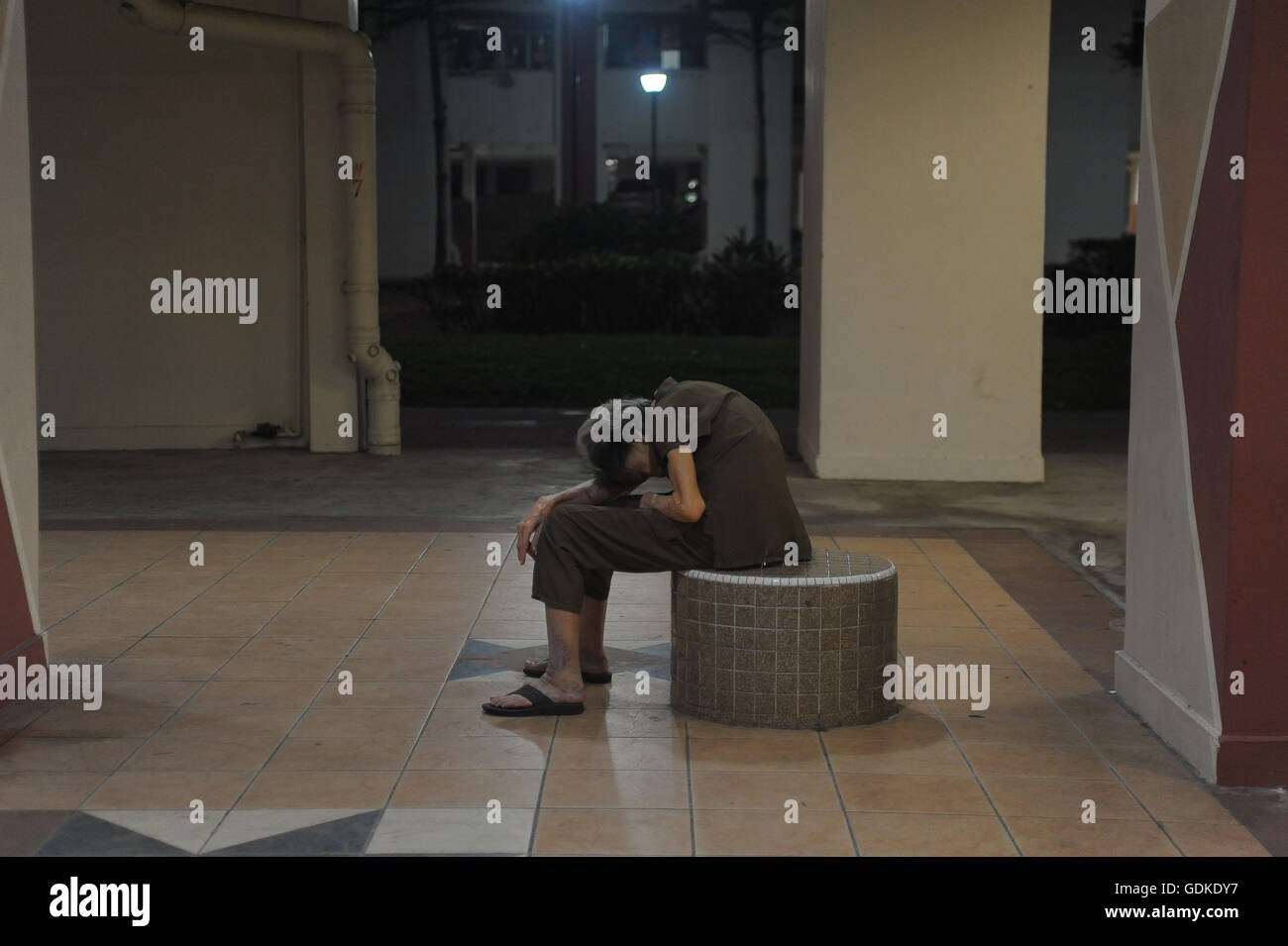 SINGAPORE, July 16, 2016. An old woman rests on a stool outside a residential housing block in the city district of Ang Mo Kio. Stock Photo