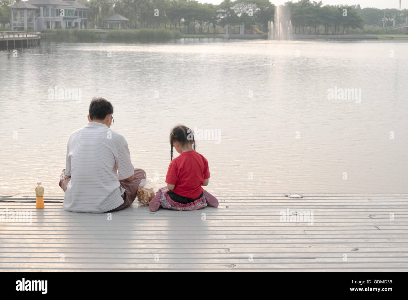 Bangkok, Thailand - March 2016: father and girl sit together feeding fish at pier on long holiday. Stock Photo