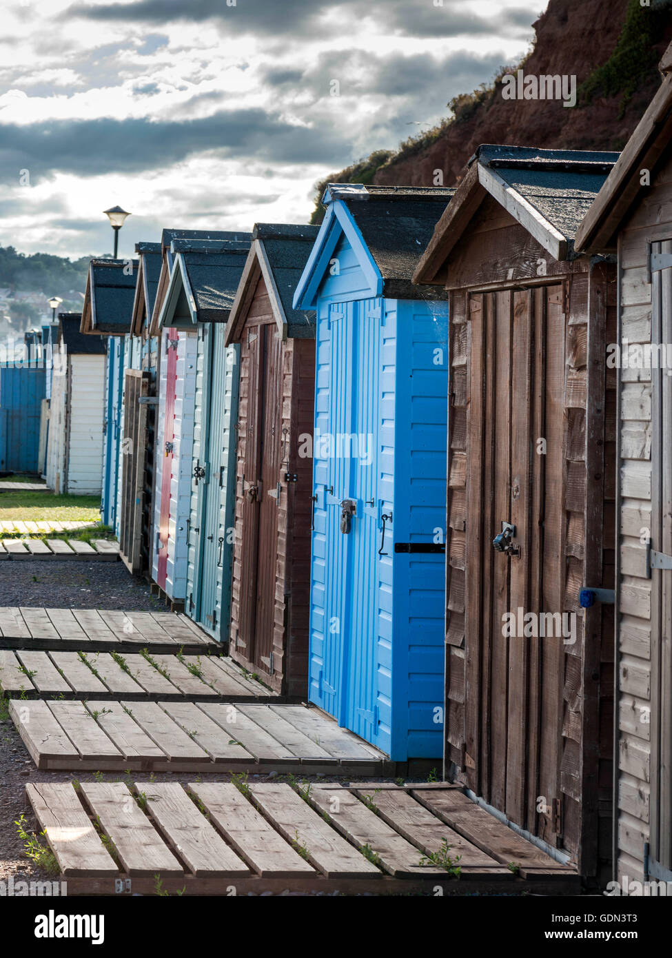 Seaside scene depicting a row of multicolored beach huts, wooden slatted pathway, pebbled beach and Jurassic coast. Stock Photo