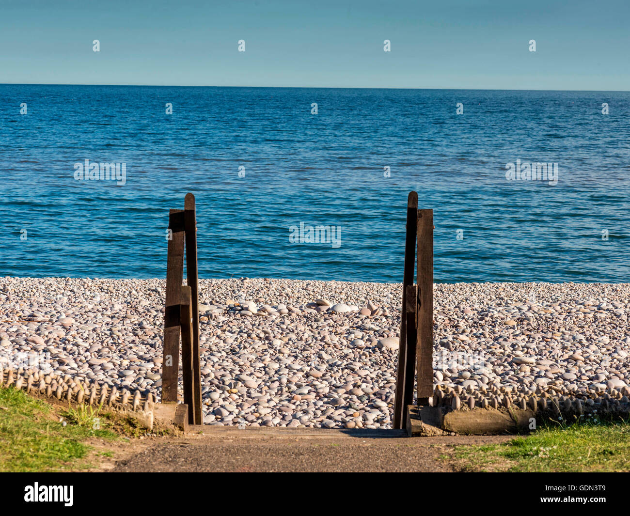 Summer seaside scene depicting pebble beach with wooden access gateway with blue ocean and sky backdrop. Stock Photo