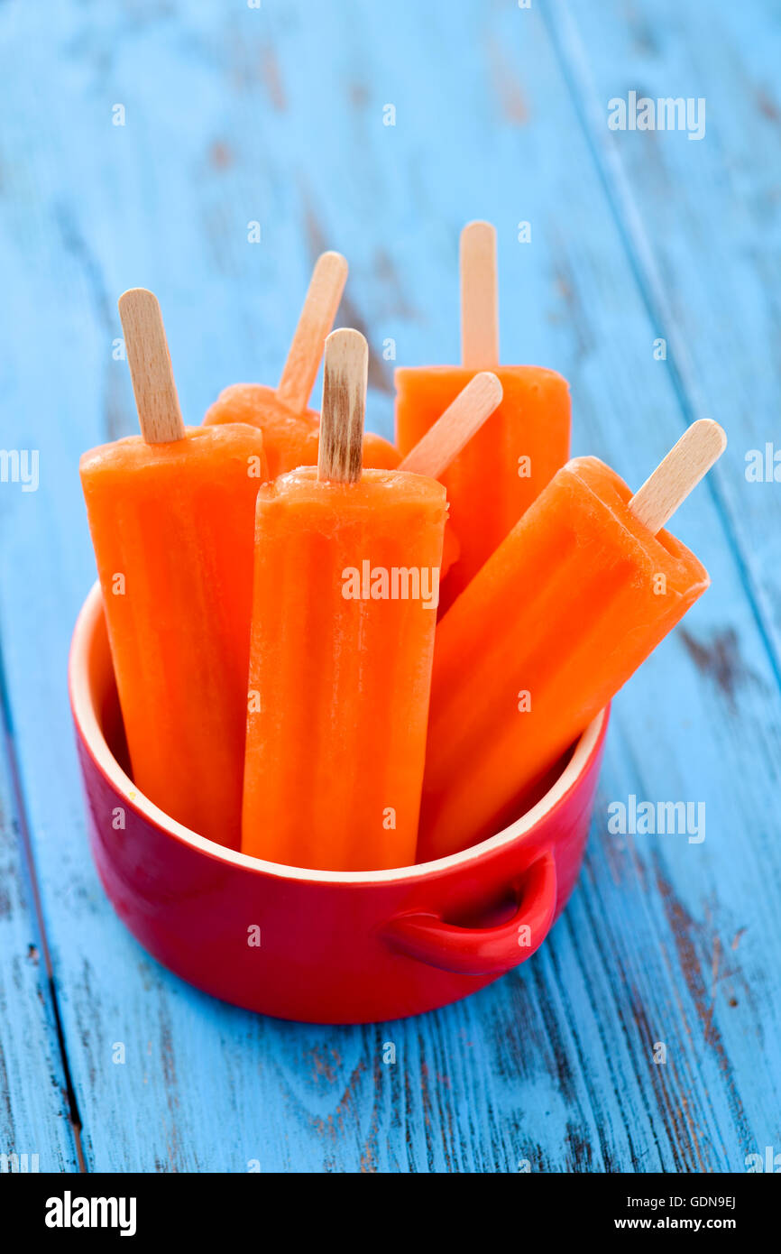 some refreshing orange flavored ice pops in a red ceramic bowl, on a blue rustic wooden table Stock Photo