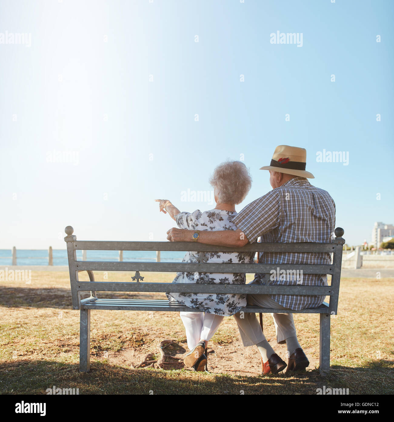 Rear view of senior couple relaxing on a bench with woman pointing out to sea. Retired man and woman sitting on a bench outdoors Stock Photo