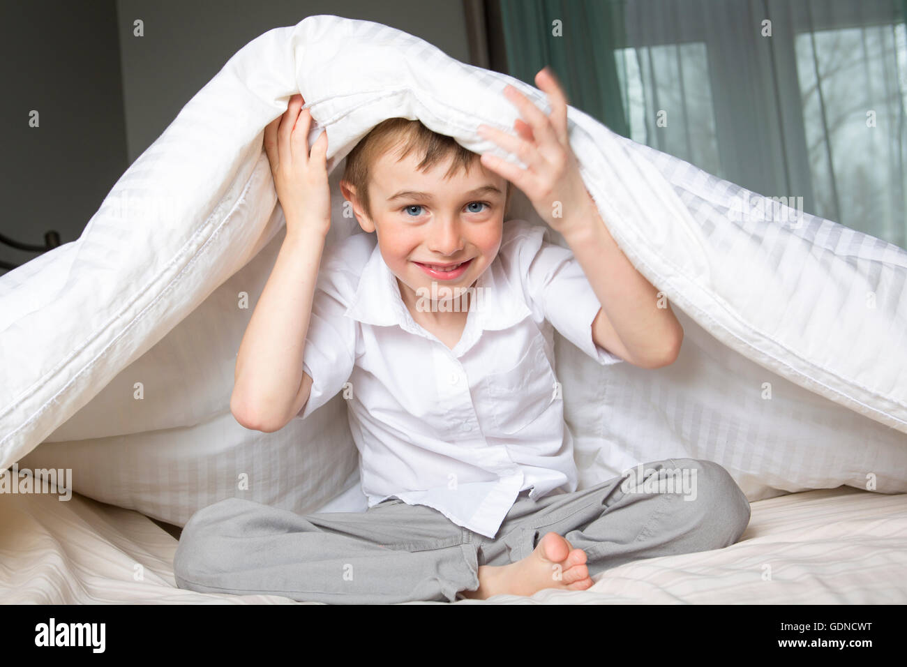 Smiling boy hiding in bed under a white blanket or coverlet Stock Photo