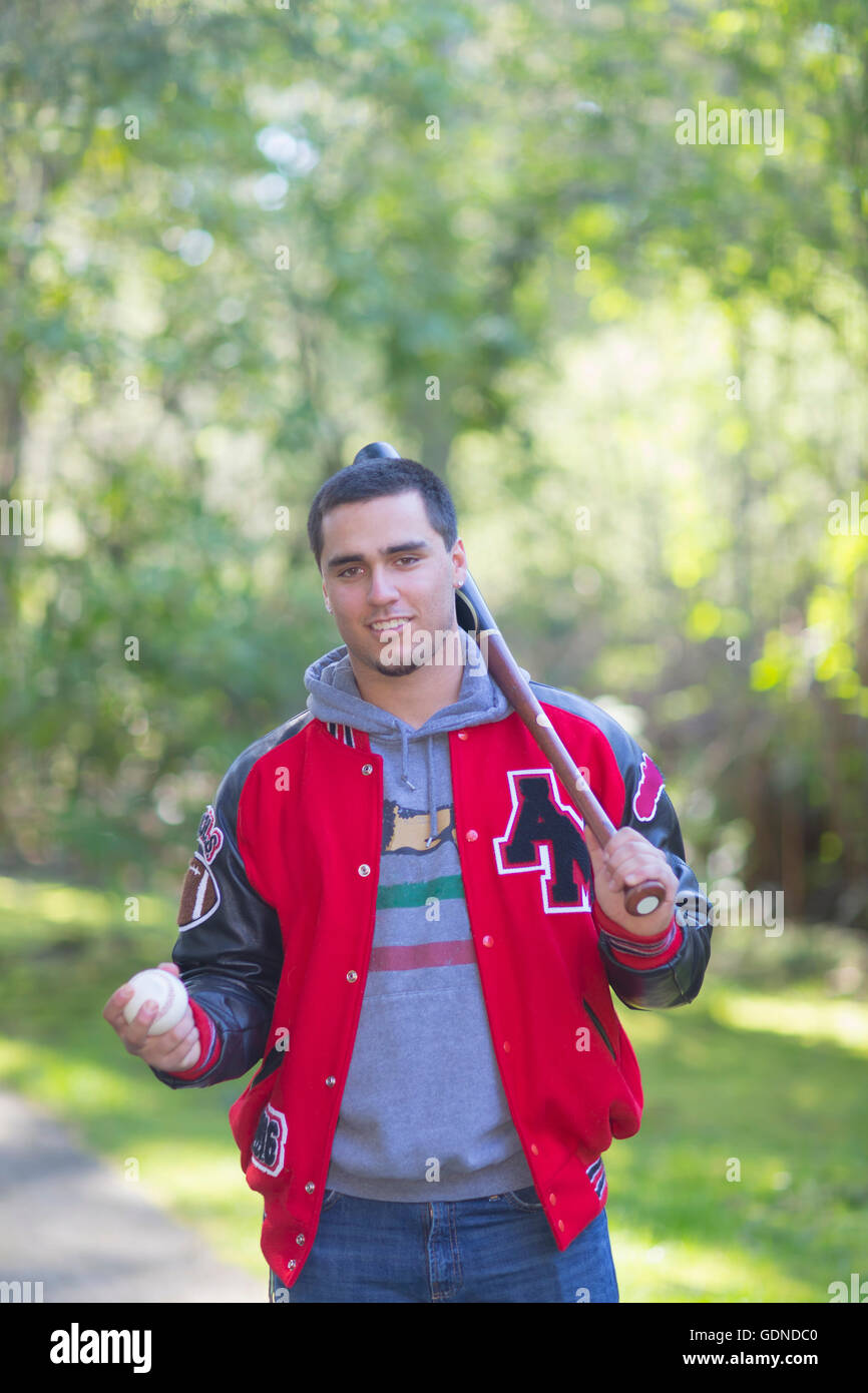 Portrait of high school senior wearing jacket, carrying baseball bat and ball in park Stock Photo