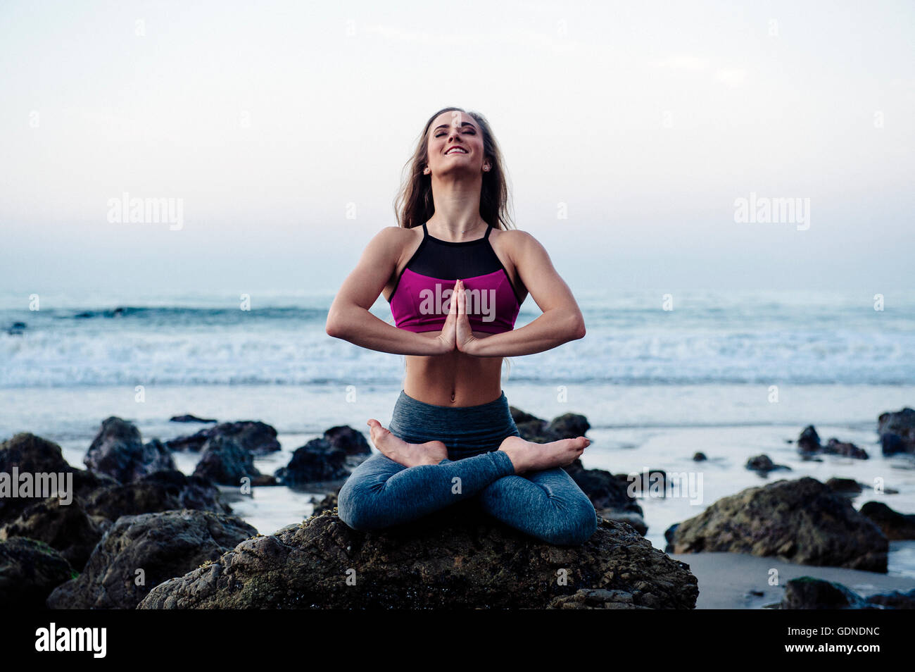 Young woman practicing lotus yoga pose on rocks at beach, Los Angeles, California, USA Stock Photo