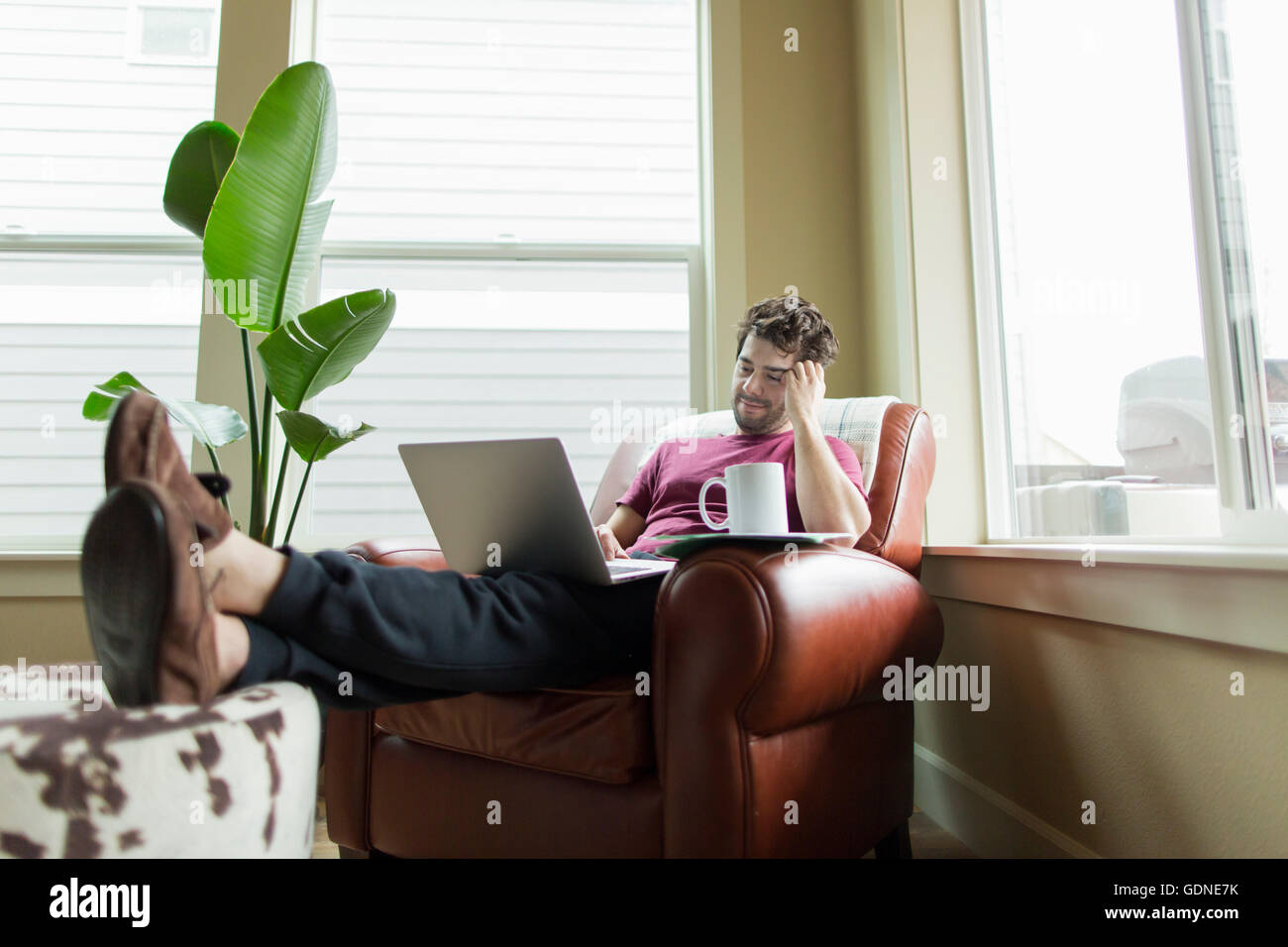 Man relaxing on living room armchair with feet up browsing laptop Stock Photo