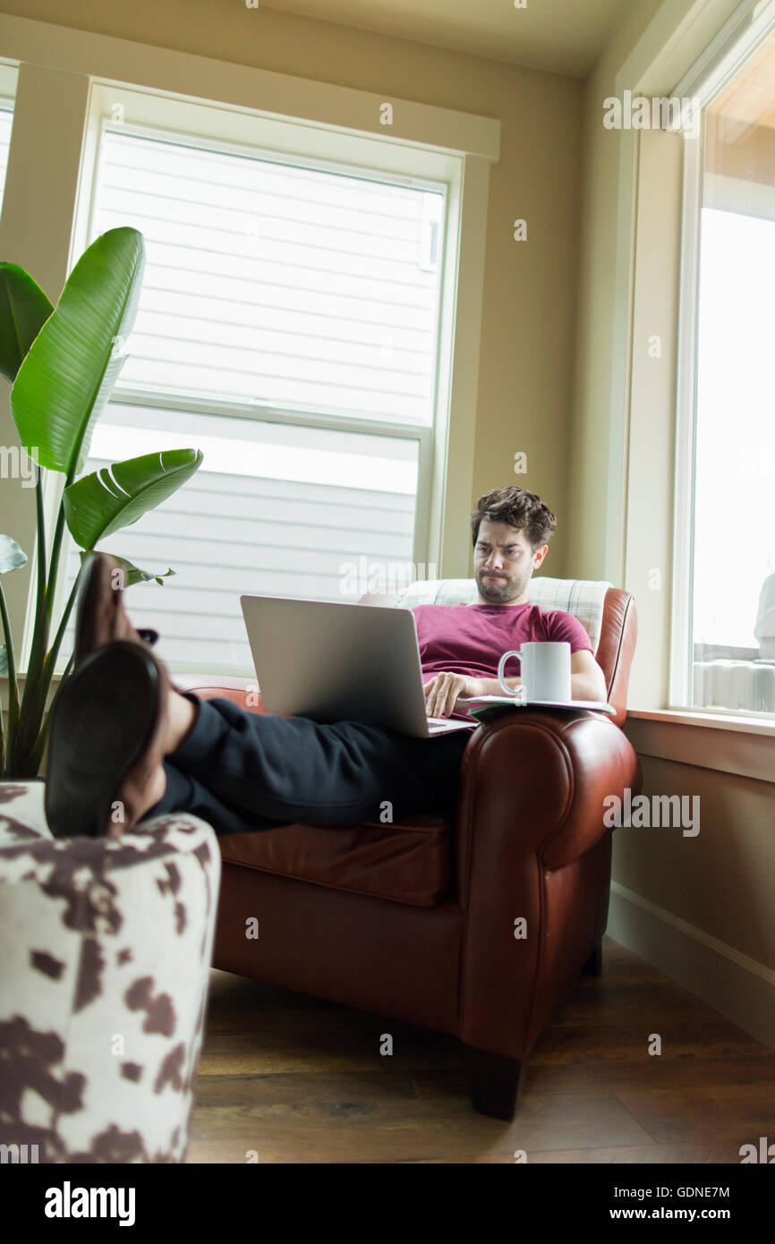 Man on living room armchair with feet up working on laptop Stock Photo